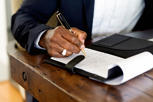 istockphoto-1323929534-170667a Close-up on mature African-American man’s hand working from home. He is wearing a navy blue jacket. He is sitting at the dinner table, white wall in the background. Horizontal close-up indoors shot with copy space. No face